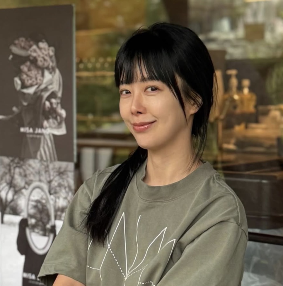 Shin Ji-soo de camiseta verde e rabo de cavalo, sorrindo de lado numa cafeteria