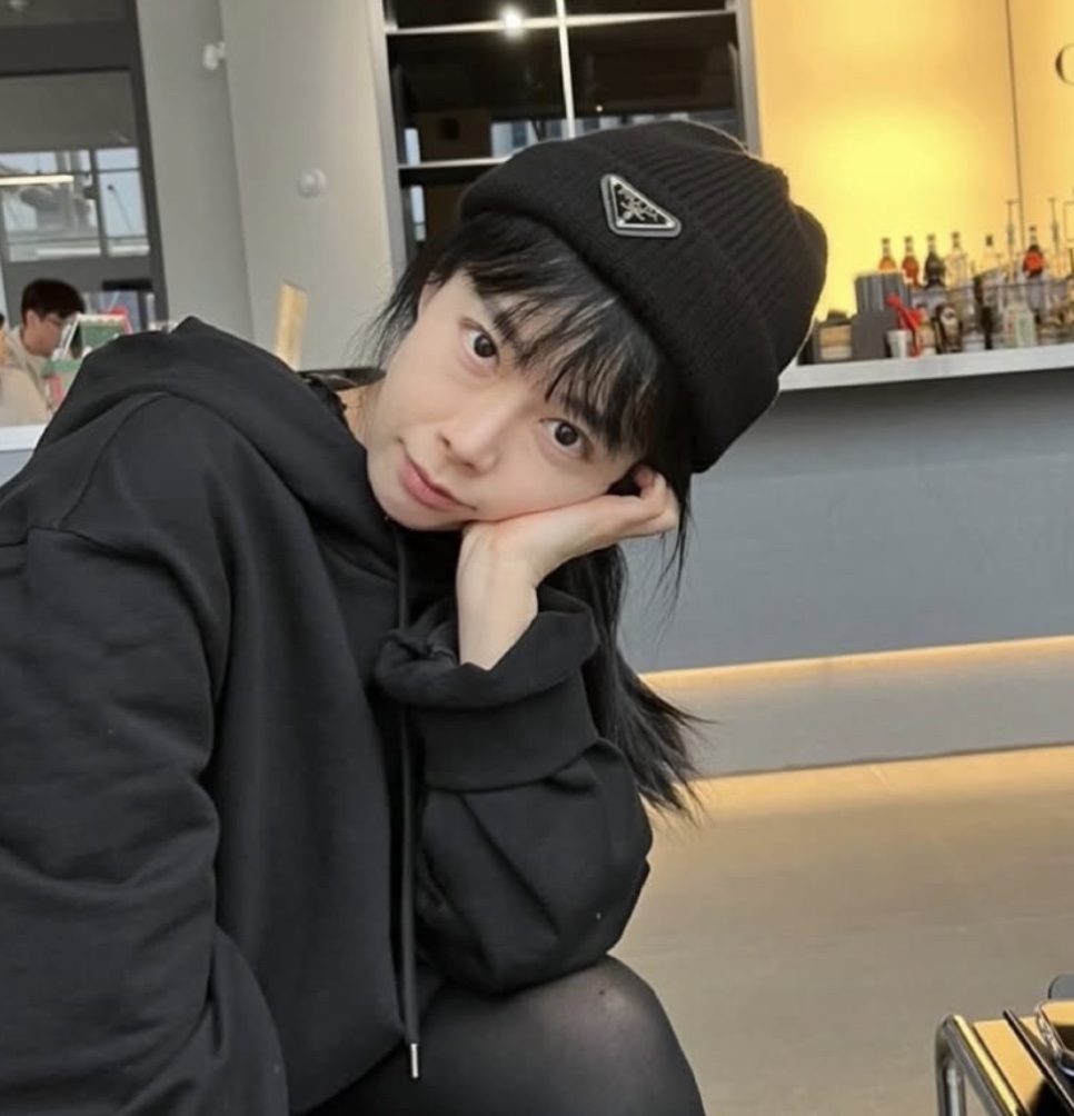 Shin Ji-soo de gorro e moletom pretos, com a mão no rosto