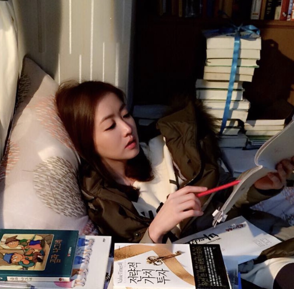 Choi Jung-won deitada numa cama cercada de pilhas de livros, lendo com uma caneta na mão