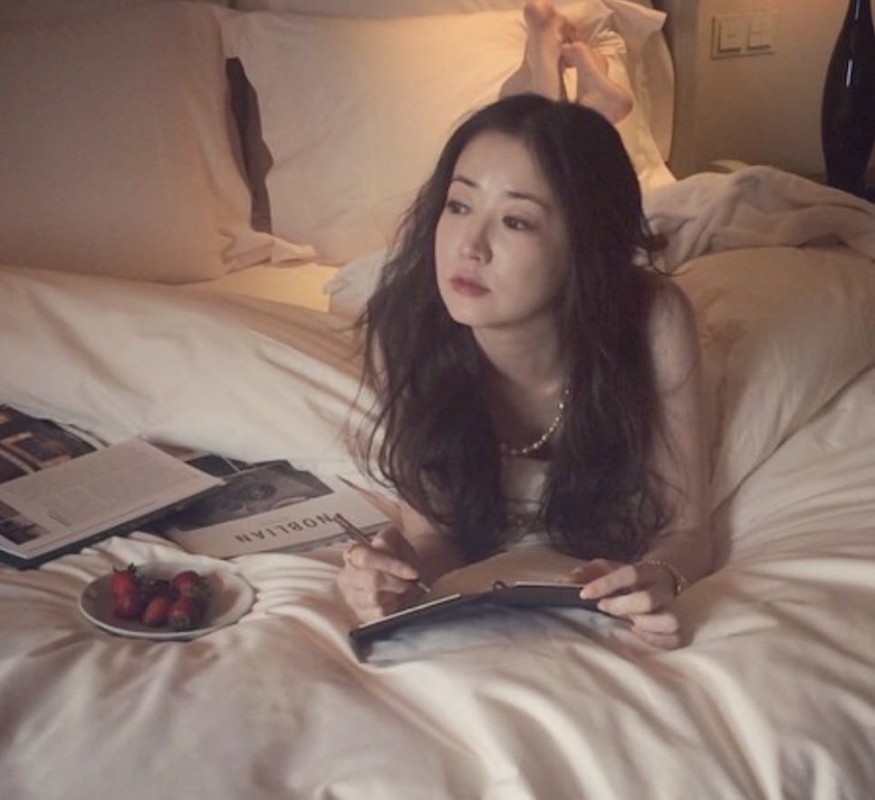 Choi Jung-won deitada de bruços numa cama branca, escrevendo num caderno