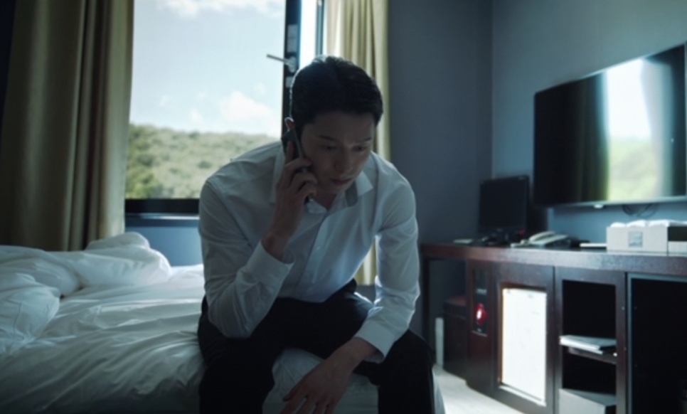 Han Jong-hyun sentado na beira da cama de um quarto de hotel, falando ao celular, em Flex X Cop 2