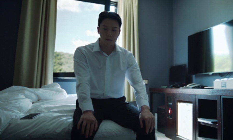 Han Jong-hyun sentado na cama de um quarto de hotel, olhando para baixo, com a janela e a TV ao fundo, em Flex X Cop 2