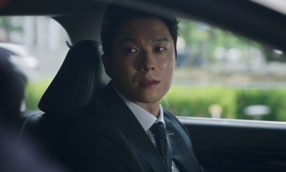 Han Jong-hyun de terno escuro, sentado no banco da frente de um carro, olhando para trás, em Flex X Cop 2