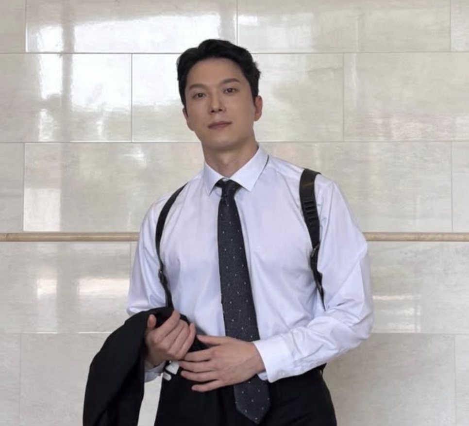 Moon Jin-seung de camisa branca, gravata e suspensórios, com o paletó na mão, diante de uma parede clara