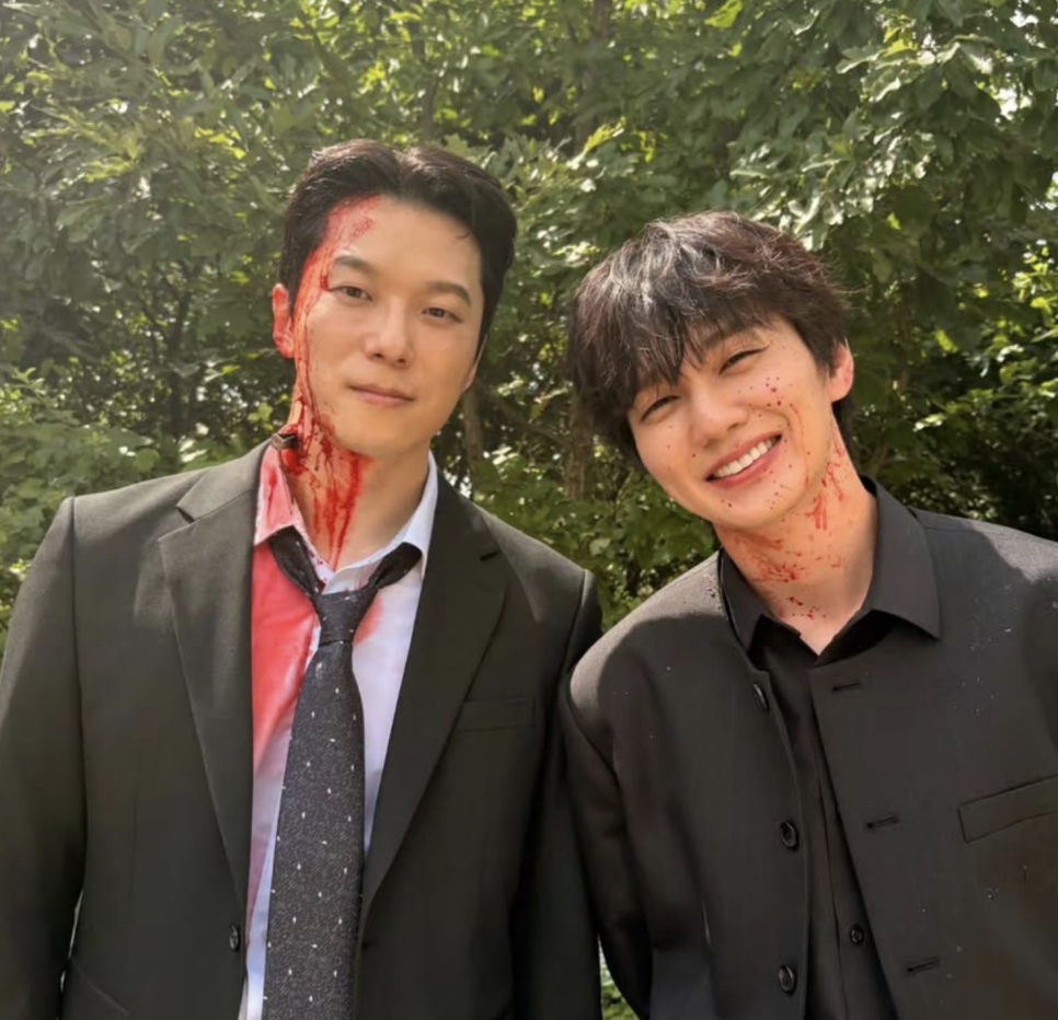 Moon Jin-seung com sangue cenográfico escorrendo pelo rosto, posando ao lado de um colega de elenco no meio de árvores