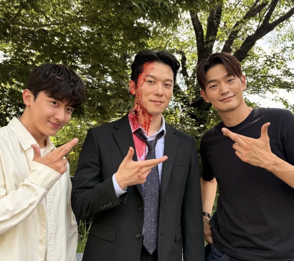 Moon Jin-seung, ainda com o sangue cenográfico, entre dois colegas de elenco fazendo sinal de V nos bastidores