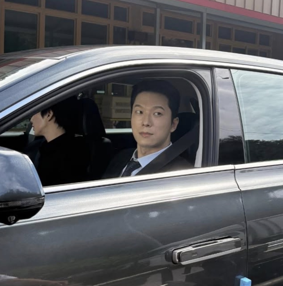 Moon Jin-seung de terno, sentado no banco do carona de um carro, olhando pela janela aberta