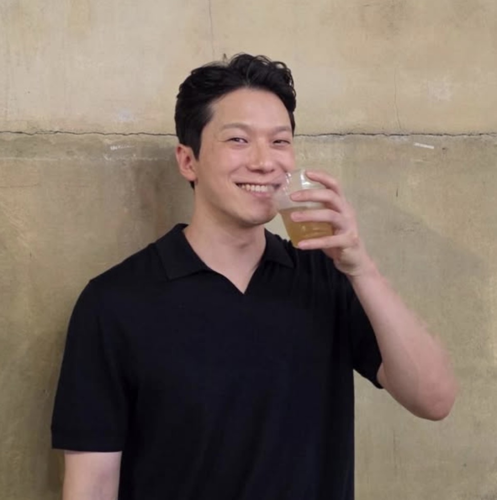 Moon Jin-seung de camisa polo preta, sorrindo com um copo na mão, diante de uma parede de concreto