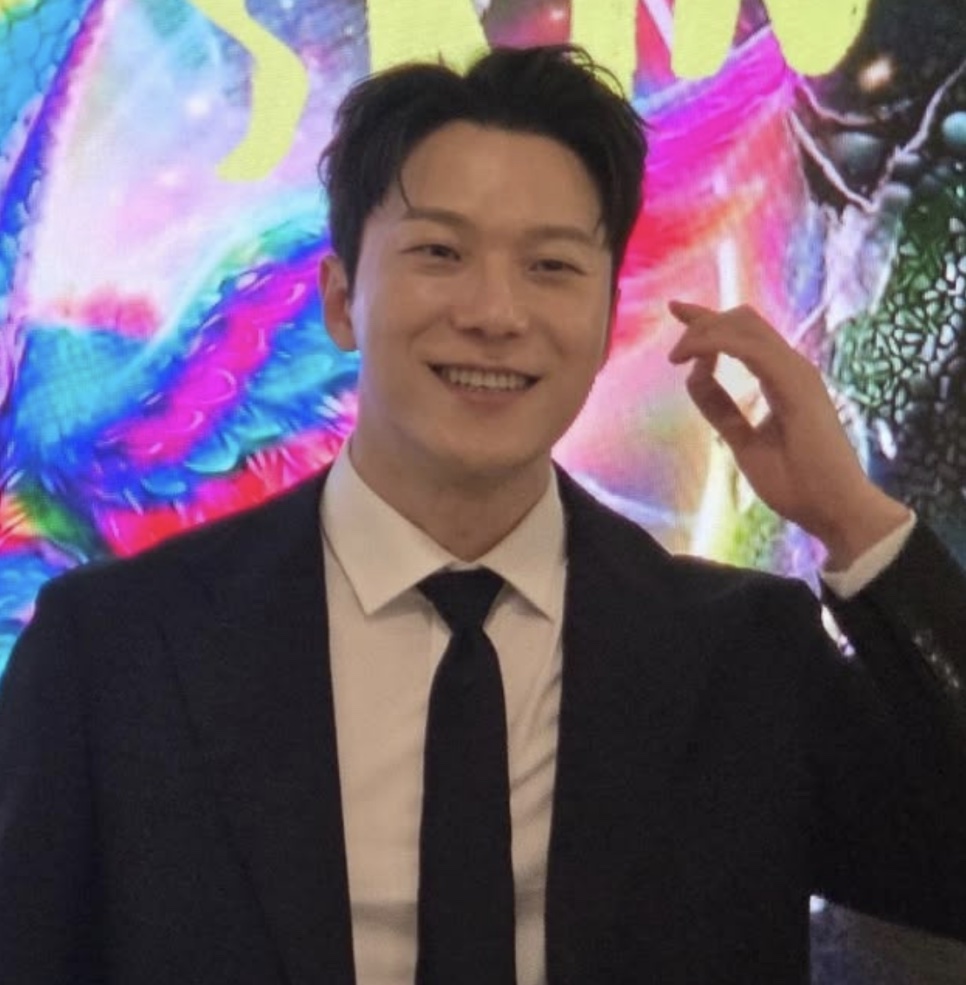 Moon Jin-seung de terno, sorrindo num evento, com um painel colorido ao fundo