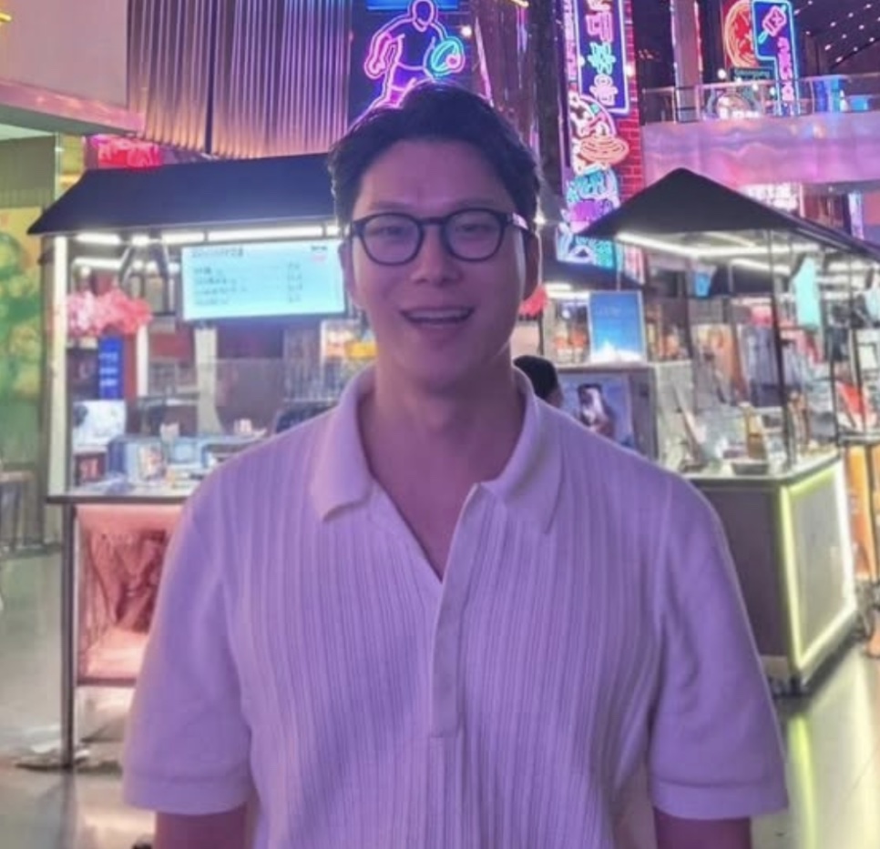 Moon Jin-seung de óculos e camisa clara, sorrindo numa galeria iluminada por letreiros de neon