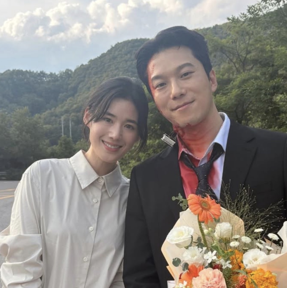 Moon Jin-seung, ainda com sangue cenográfico no rosto, segurando um buquê ao lado de uma colega de elenco, com montanhas ao fundo