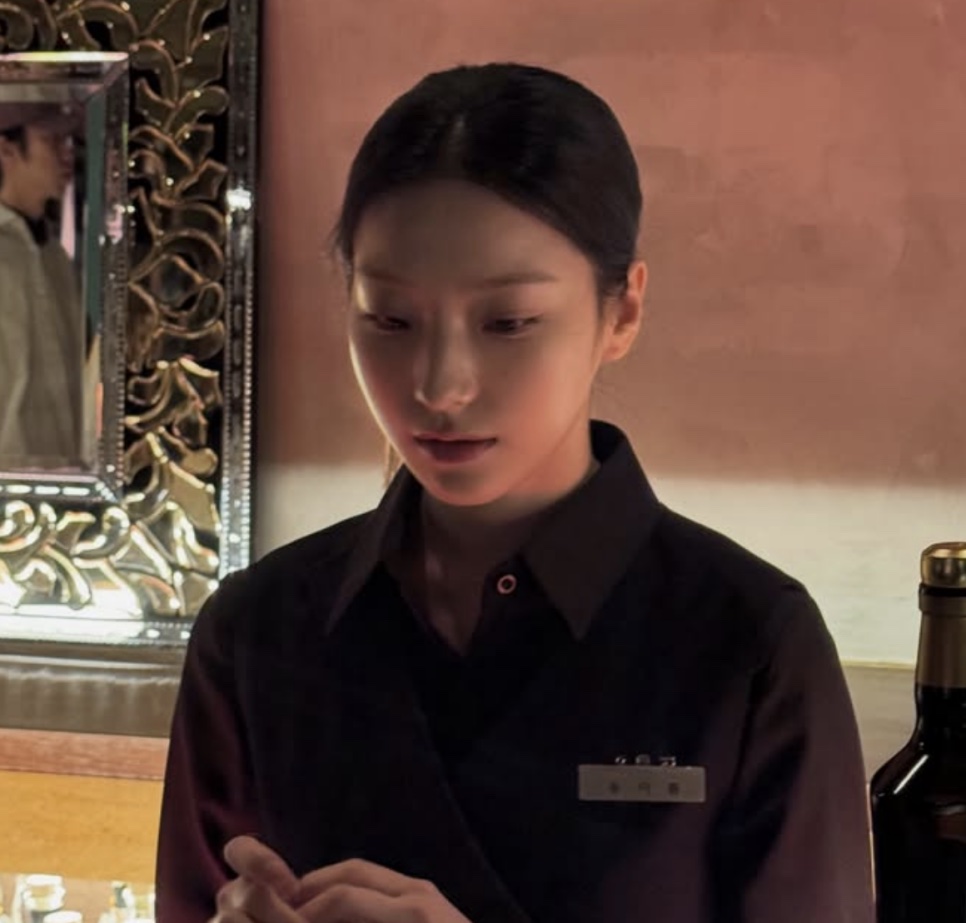 Lee Sae-rom de uniforme preto e crachá, olhando para baixo, no cenário do bar