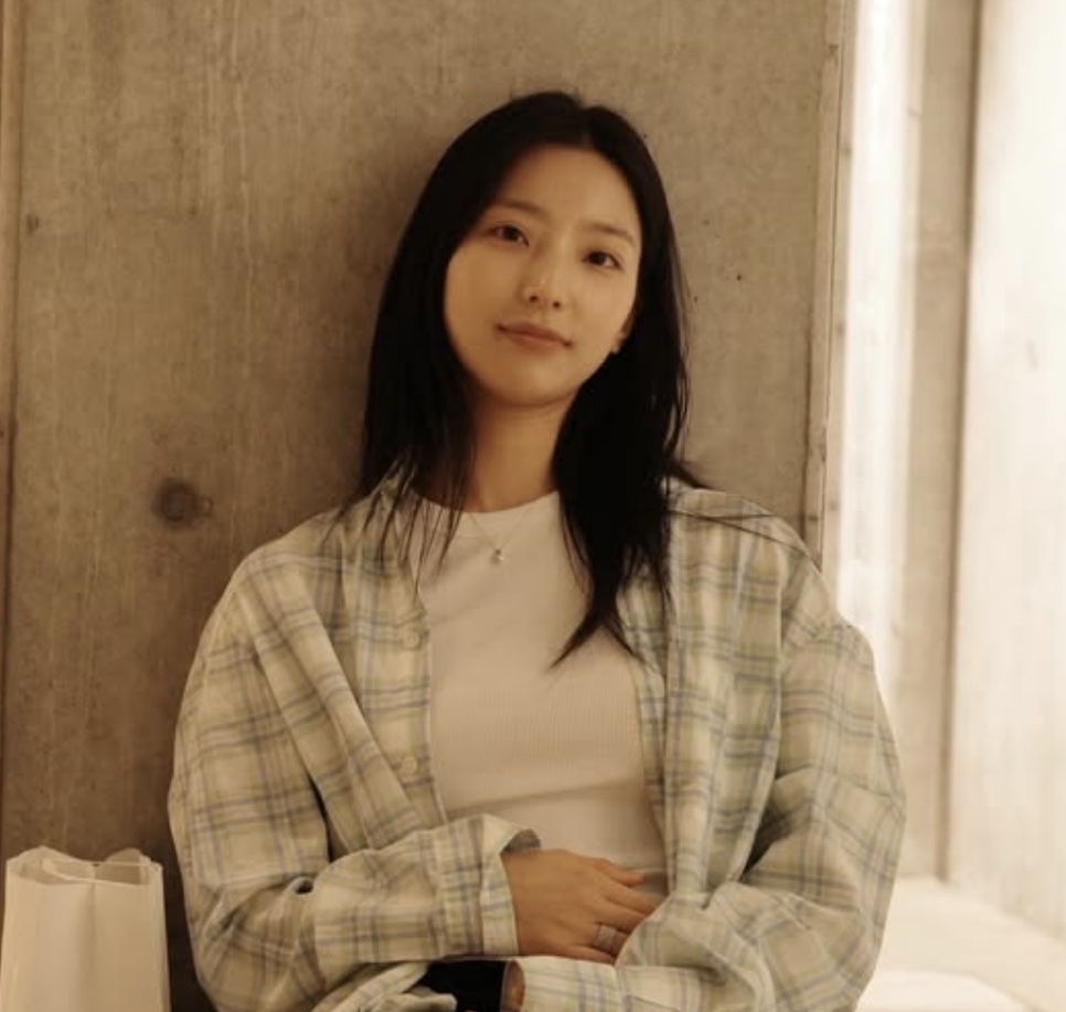 Lee Sae-rom de camisa xadrez sobre camiseta branca, sentada encostada numa parede