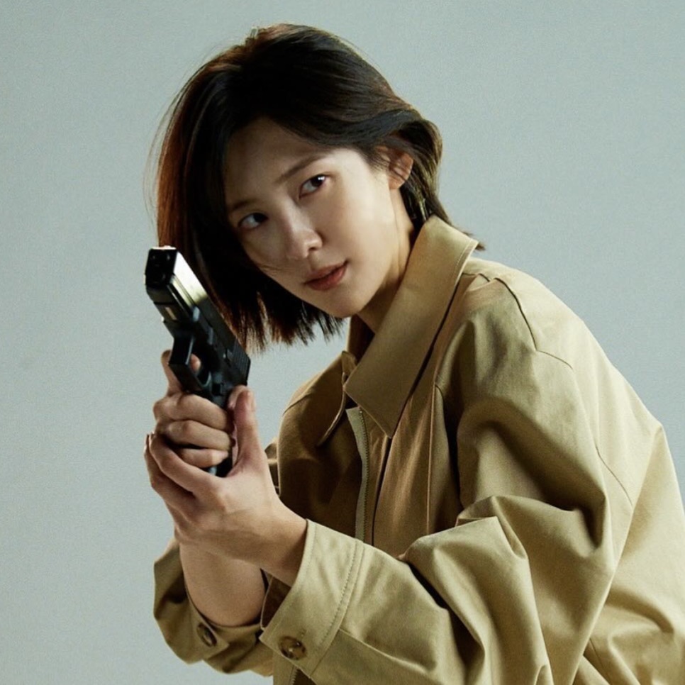 Park Ji-hyun de trench coat bege segurando uma pistola, olhando para a câmera