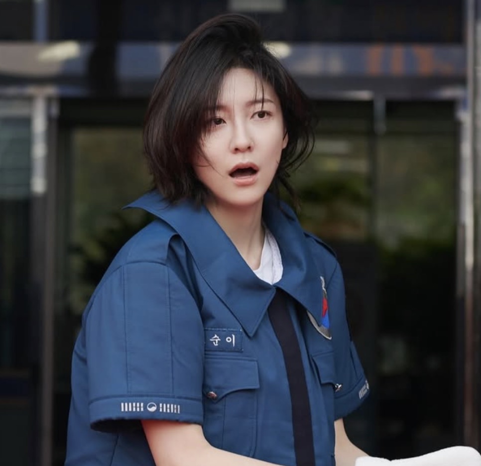 Park Ji-hyun de uniforme policial azul, com expressão de susto, na porta da delegacia
