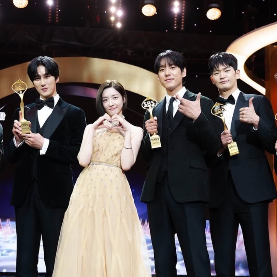 Park Ji-hyun de vestido dourado entre três atores de smoking, todos segurando troféus no palco