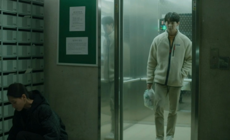 Woo-jae andando pelo corredor do prédio com um saco na mão, visto de longe