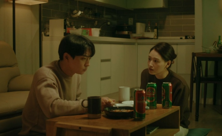 Woo-jae e Ji-su sentados à mesa de casa, com comida e latas de bebida