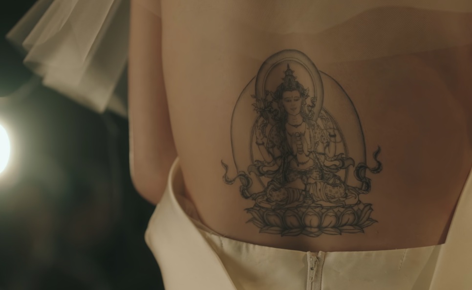 Close das costas da noiva com o vestido aberto, mostrando a tatuagem de uma figura budista sentada sobre uma flor de lótus