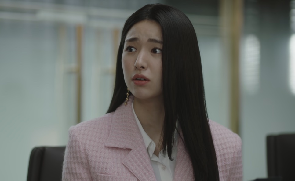 Kim Hwa-young de blazer rosa, sentada em uma sala de reunião envidraçada, com expressão abatida