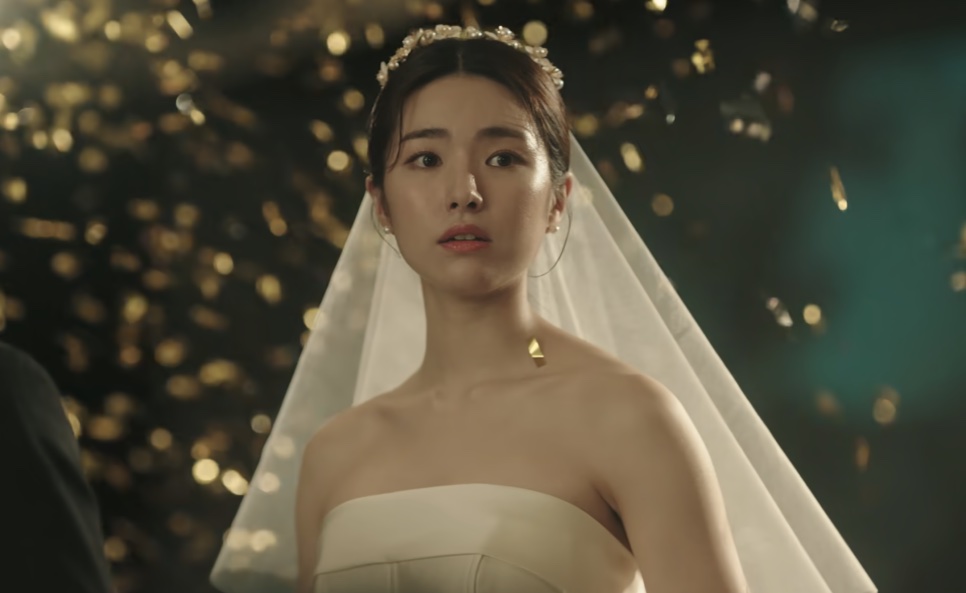 A noiva Kim Hwa-young de véu e vestido claro, em pé diante de uma parede de luzes douradas