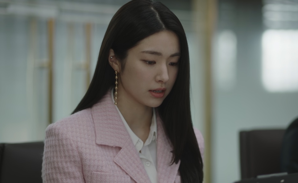 Kim Hwa-young de blazer rosa e brinco comprido, olhando para baixo em uma sala de escritório