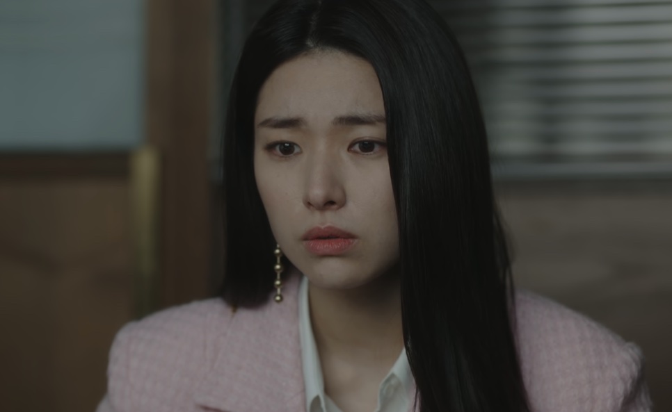Kim Hwa-young de blazer rosa, com os olhos marejados, diante de uma janela com persiana