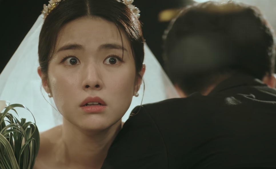 A noiva Kim Hwa-young de véu, abraçada a um homem de terno, com o olhar perdido