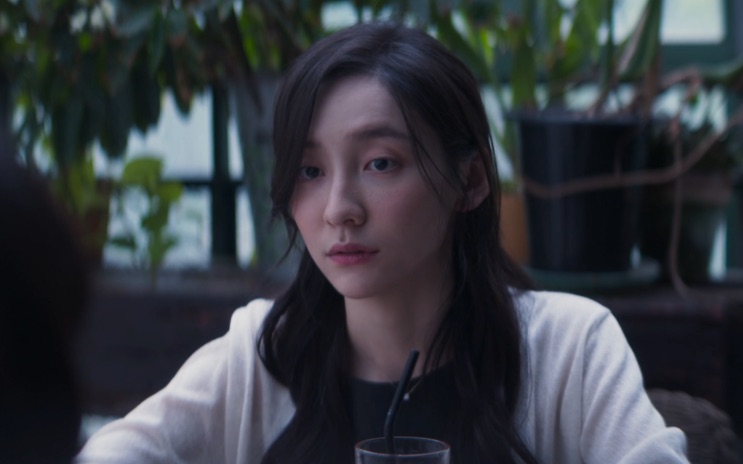 Park Ji-hyun de cardigã claro, sentada a uma mesa cercada de plantas
