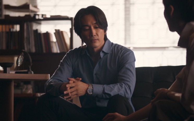 Song Seung-heon de camisa azul, sentado num sofá de frente para outra pessoa