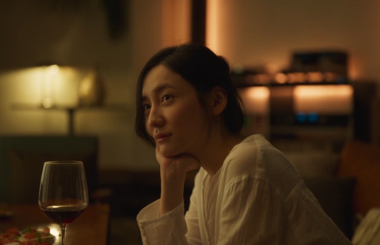 Cho Yeo-jeong de blusa branca, apoiada na mão, com uma taça de vinho tinto na mesa