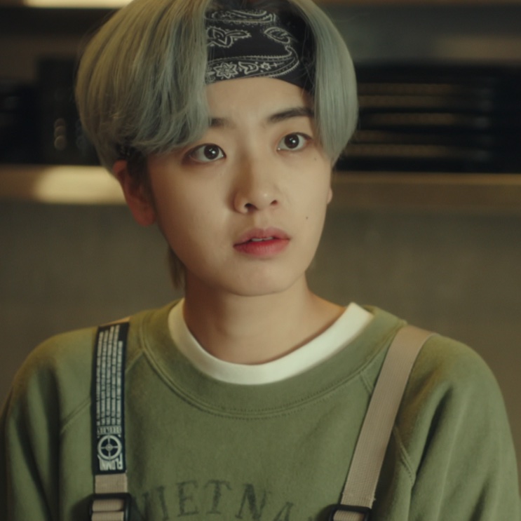 Ma Hyun-yi de bandana preta e cabelo curto acinzentado, de moletom verde com avental, na cozinha do Danbam