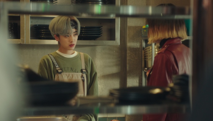 Ma Hyun-yi de avental na cozinha do Danbam, conversando com uma pessoa de jaqueta vermelha