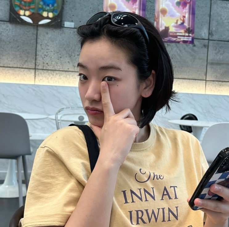 Lee Joo-young de camiseta amarela e óculos de sol na cabeça, sentada em uma praça de alimentação