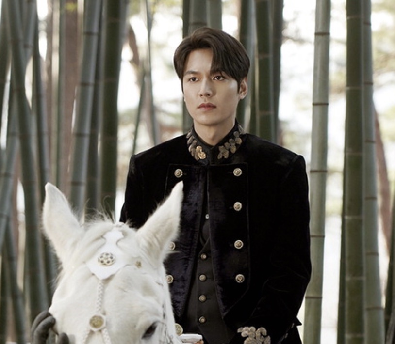 Lee Min-ho de uniforme escuro ao lado de um cavalo branco em um bambuzal, em O Rei Eterno