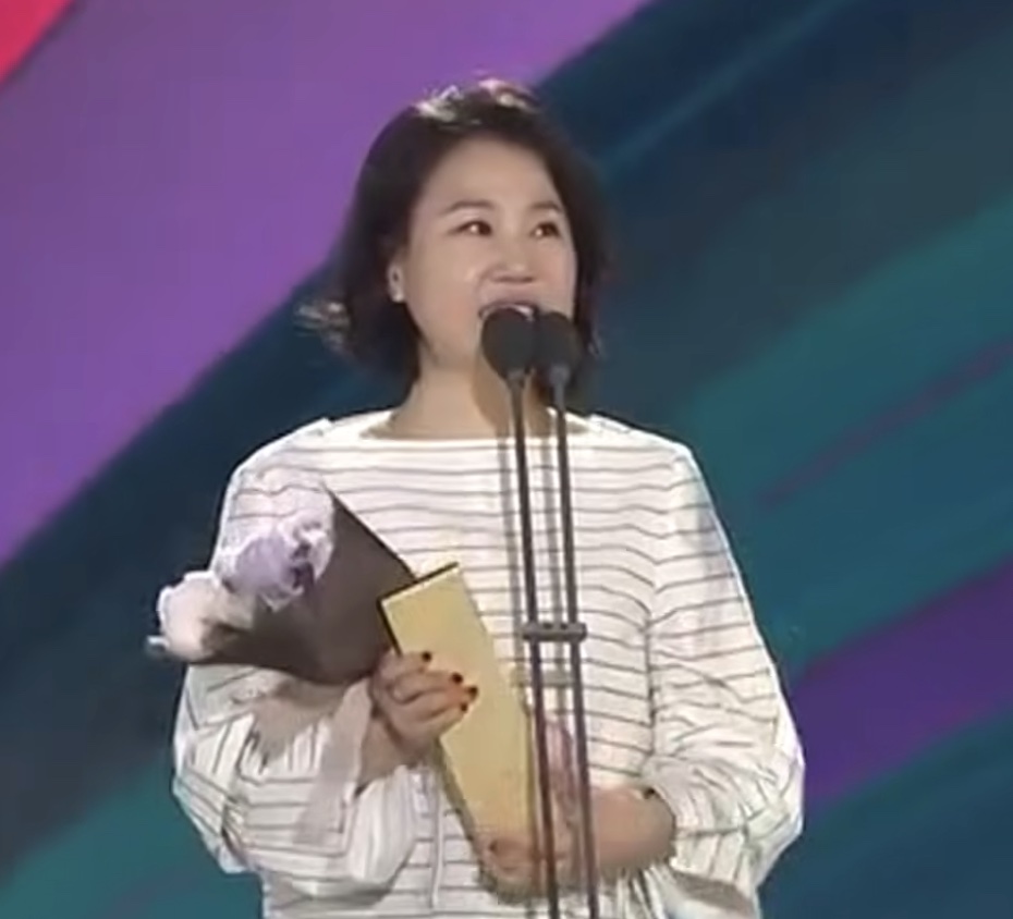 Kim Eun-sook no palco de uma premiação, segurando um troféu e um buquê de flores