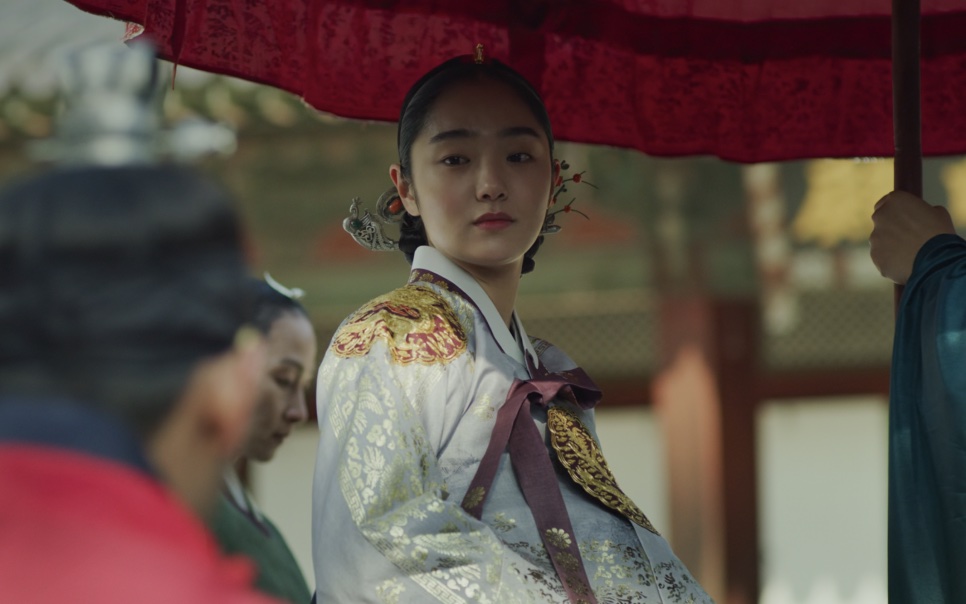 Kim Hye-jun de hanbok branco e dourado, sob uma sombrinha vermelha, no papel da rainha de Kingdom