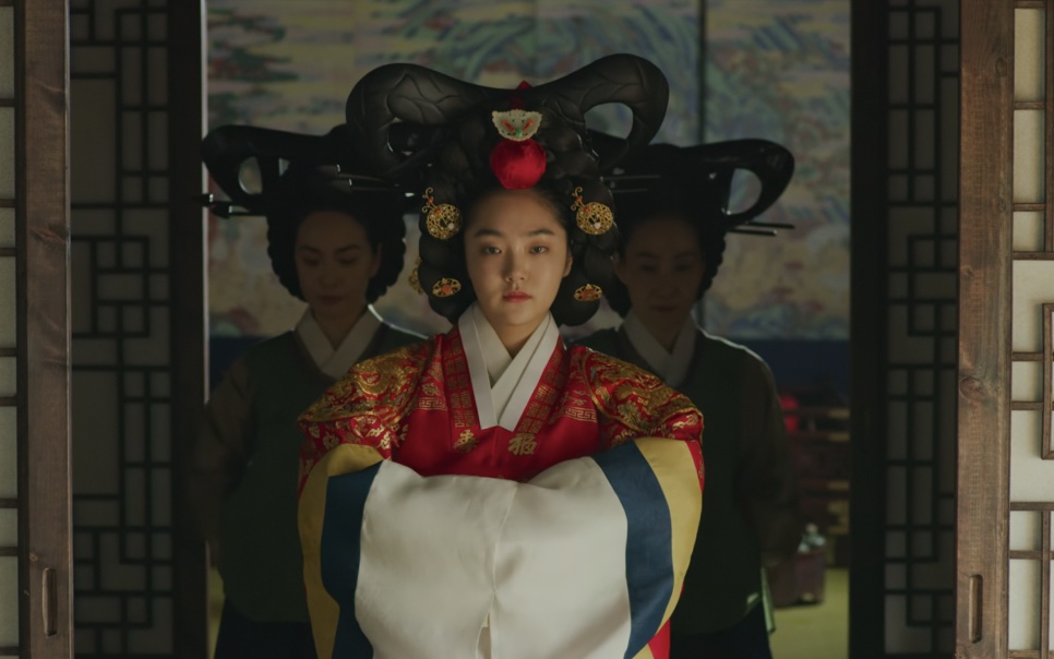 A rainha de Kingdom entrando no salão com o penteado cerimonial alto, seguida pelas damas da corte