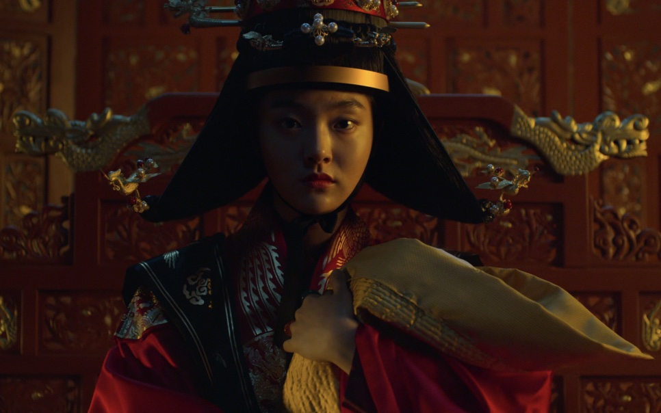 A rainha de Kingdom no trono, em uma cena escura, de chapéu preto e roupa vermelha