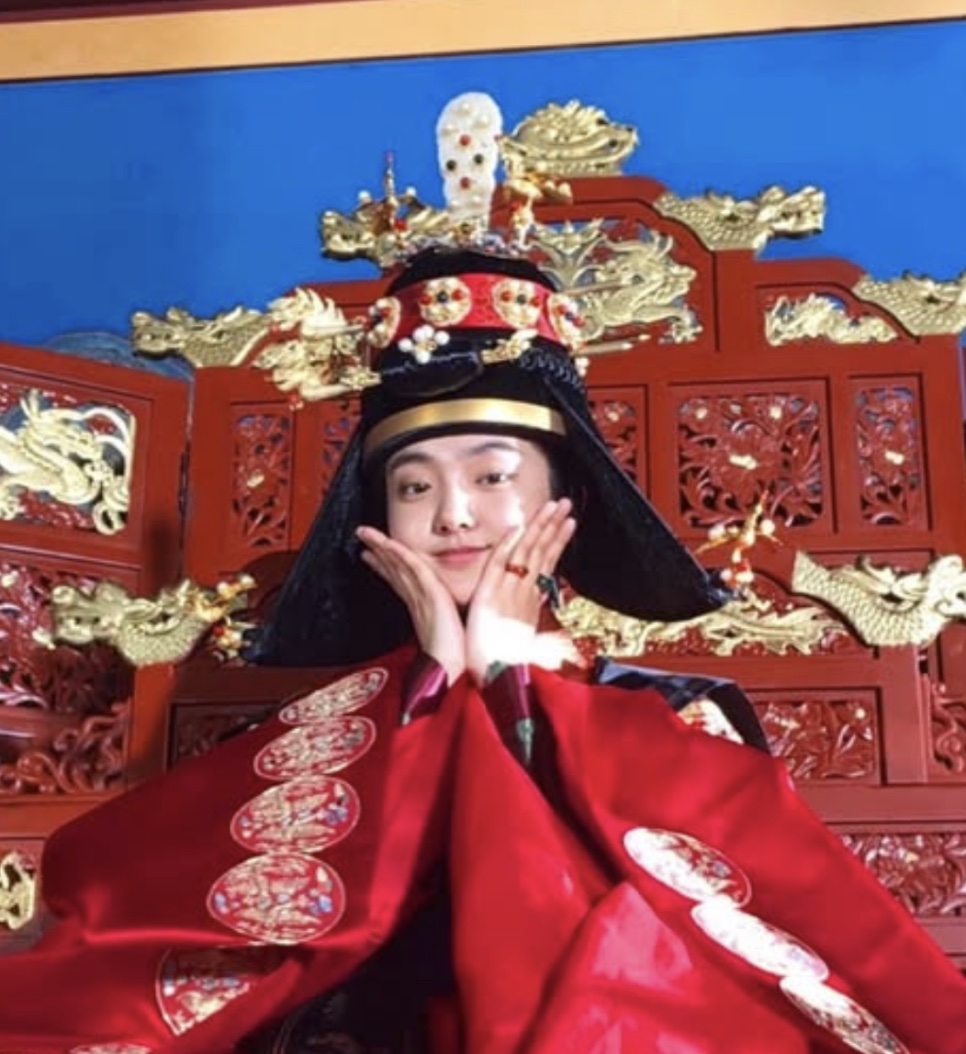 Kim Hye-jun fazendo pose com as mãos no rosto, sentada no trono com o figurino da rainha, nos bastidores de Kingdom