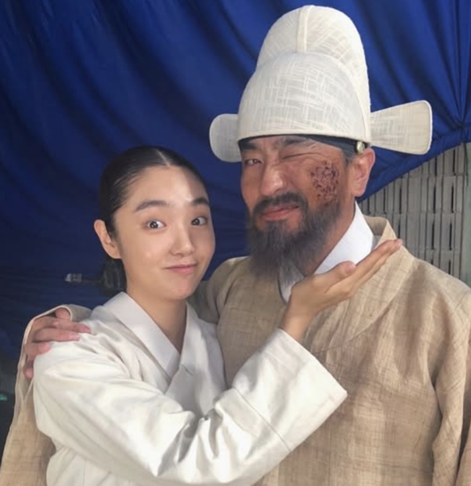 Kim Hye-jun de hanbok branco abraçando um ator do elenco com maquiagem de ferimentos, nos bastidores de Kingdom
