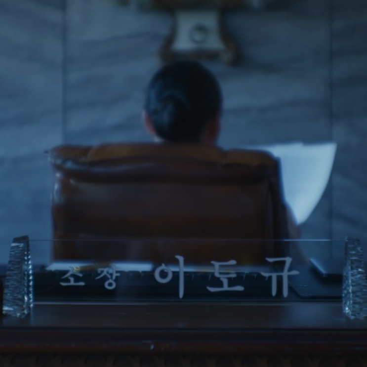 Placa de mesa com o nome do diretor Lee Do-gyu e, atrás dela, uma cadeira de escritório de costas