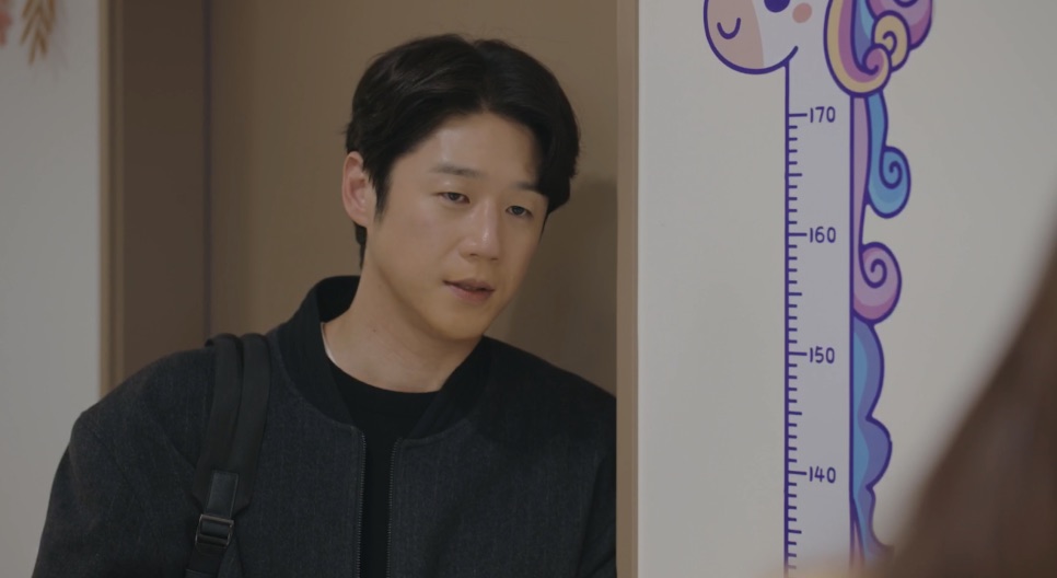 Jung Joon-won de jaqueta escura e mochila, numa sala com régua de altura infantil na parede