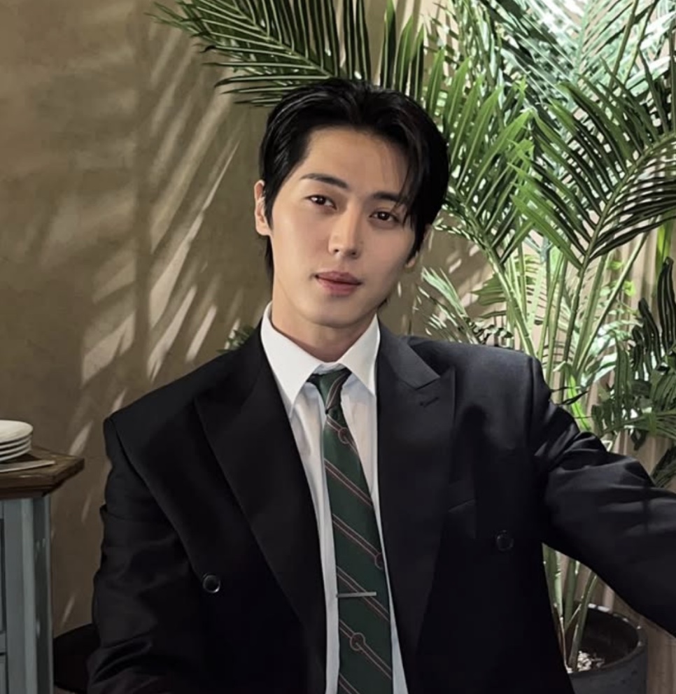 Lee Joo-ahn de terno preto e gravata verde listrada, sentado na frente de uma palmeira