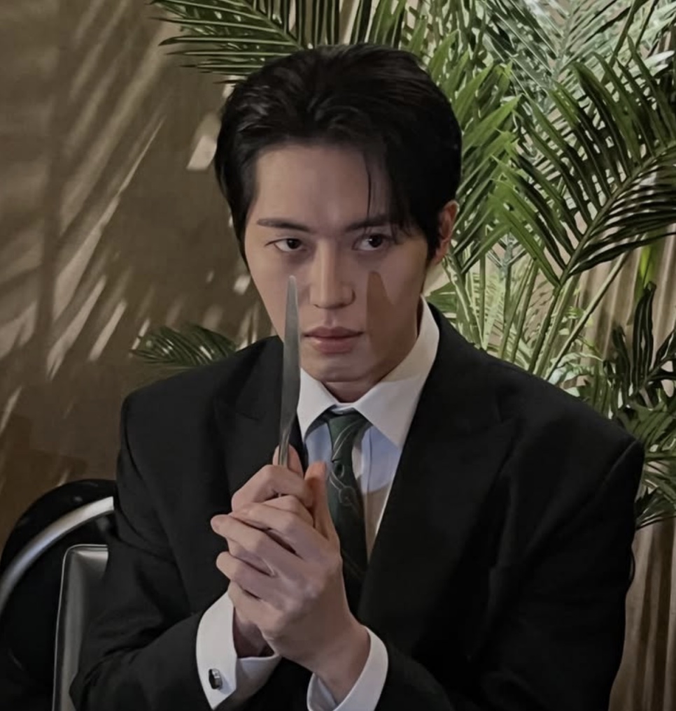 Lee Joo-ahn de terno preto, olhando sério pra câmera e segurando uma faca de mesa na frente do rosto