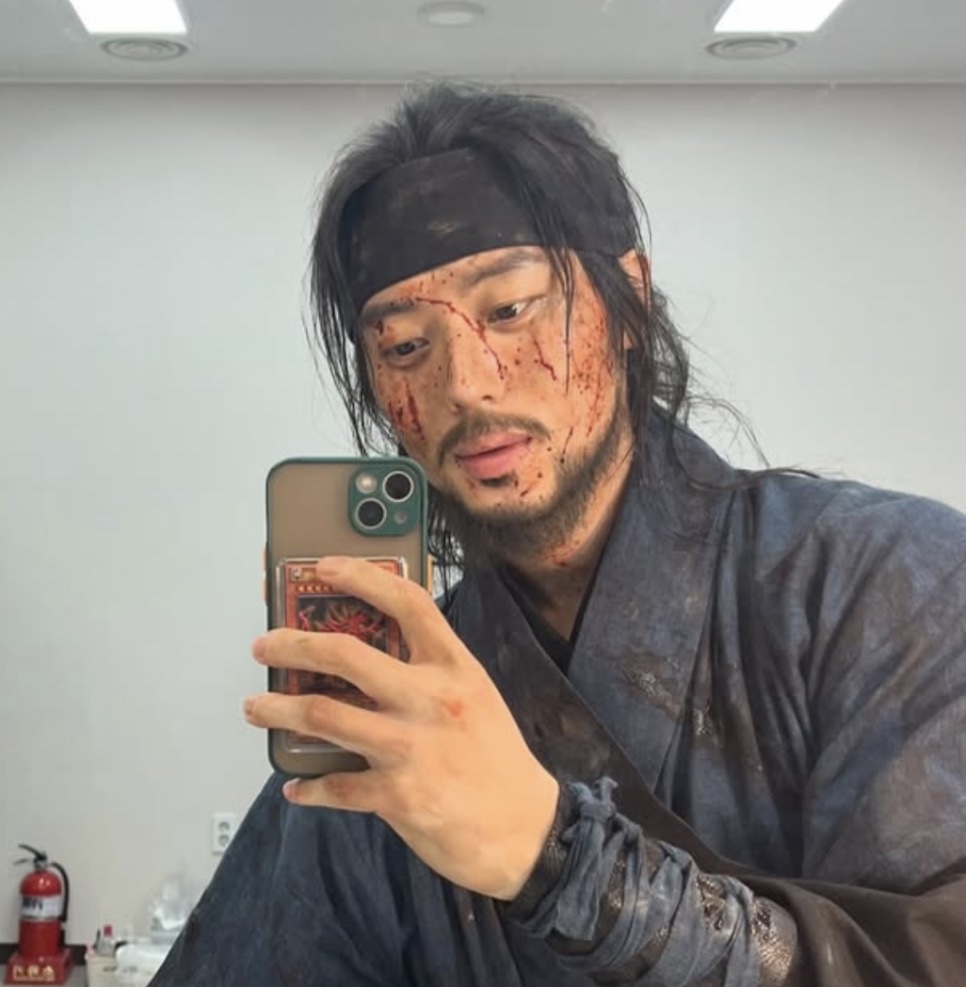 Lee Joo-ahn caracterizado de Gong-gil, com faixa na cabeça e maquiagem de sangue no rosto, tirando selfie no espelho