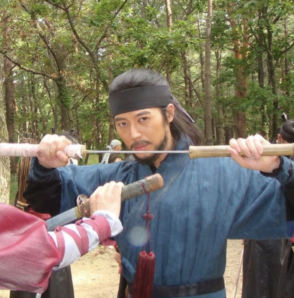Lee Joo-ahn de Gong-gil, com roupa tradicional azul e faixa preta na cabeça, cruzando duas espadas numa floresta