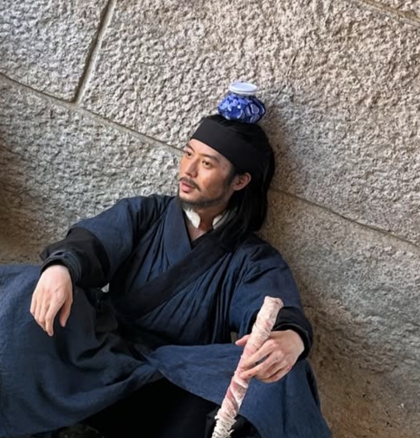 Lee Joo-ahn de Gong-gil, sentado encostado numa parede de pedra com uma tigela de porcelana azul equilibrada na cabeça