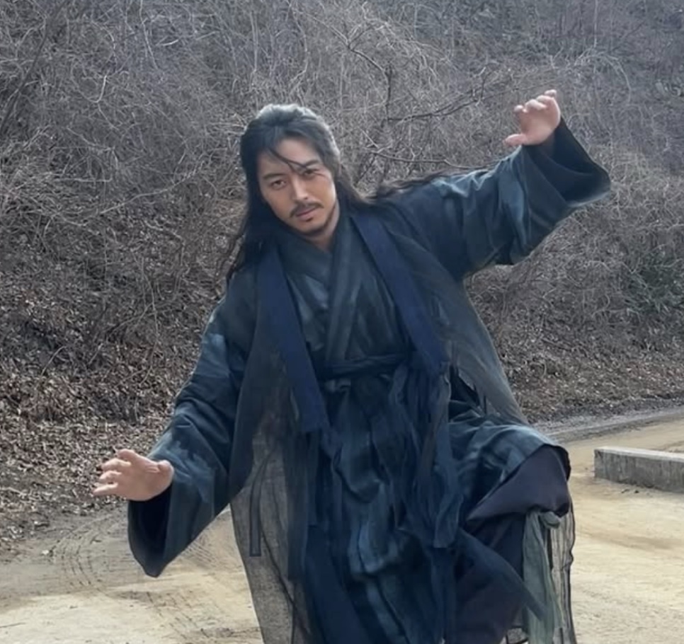 Lee Joo-ahn de Gong-gil, de roupa tradicional escura e cabelo comprido, fazendo uma pose de dança numa estrada de terra