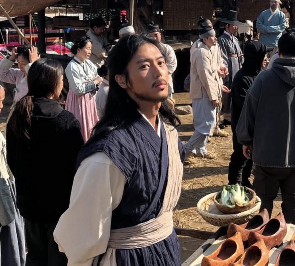 Lee Joo-ahn de Gong-gil no meio da equipe e dos figurantes, num cenário de mercado antigo